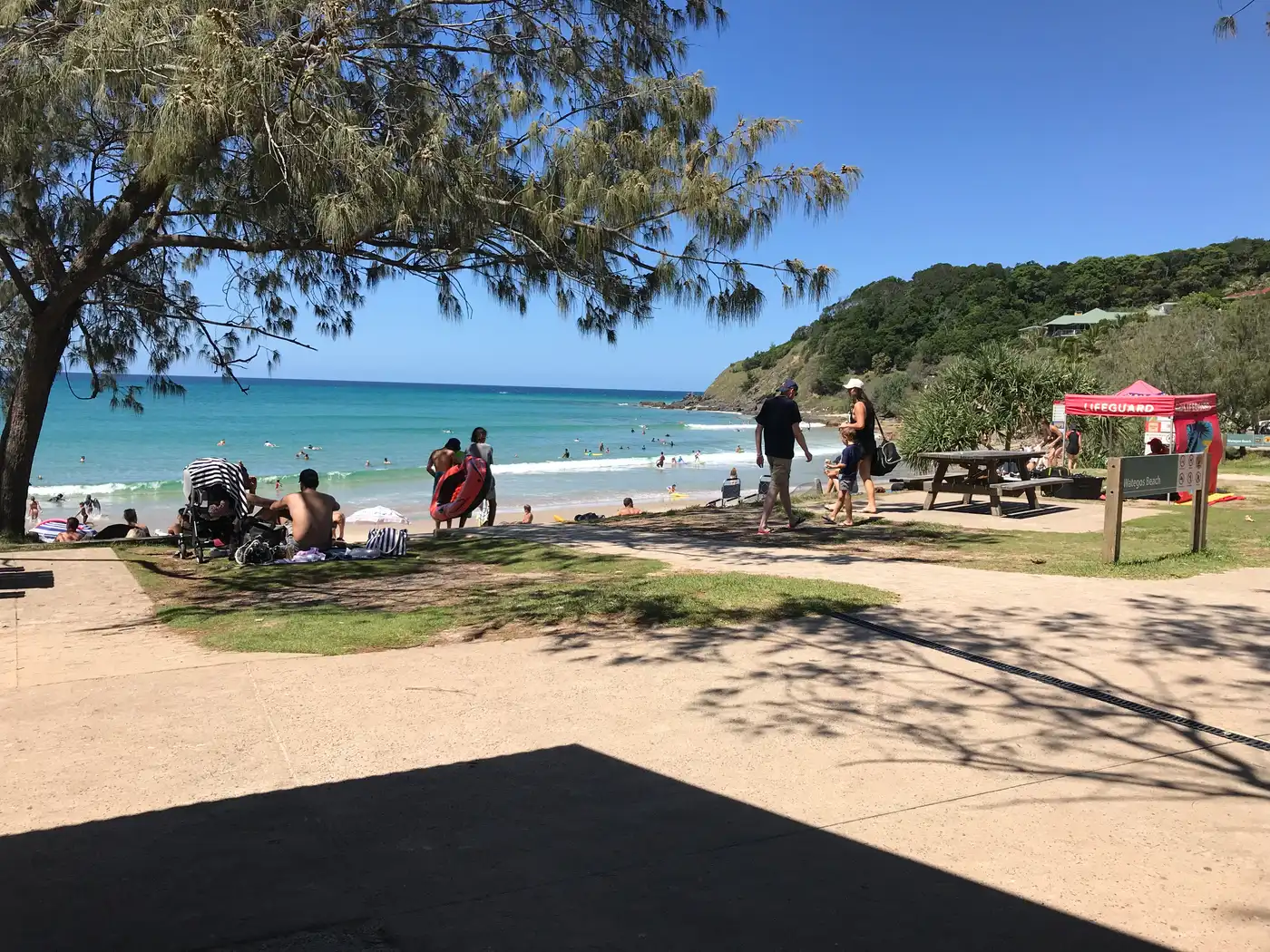 Beachgoers and a red lifeguard tent on a sunny day at a Byron Bay beach with the headland behind