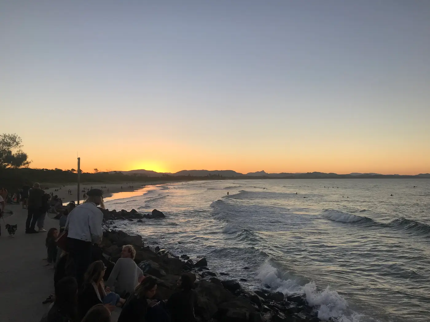 Crowd gathered on the rocky breakwall at Byron Bay watching the sun set over the headland and ocean